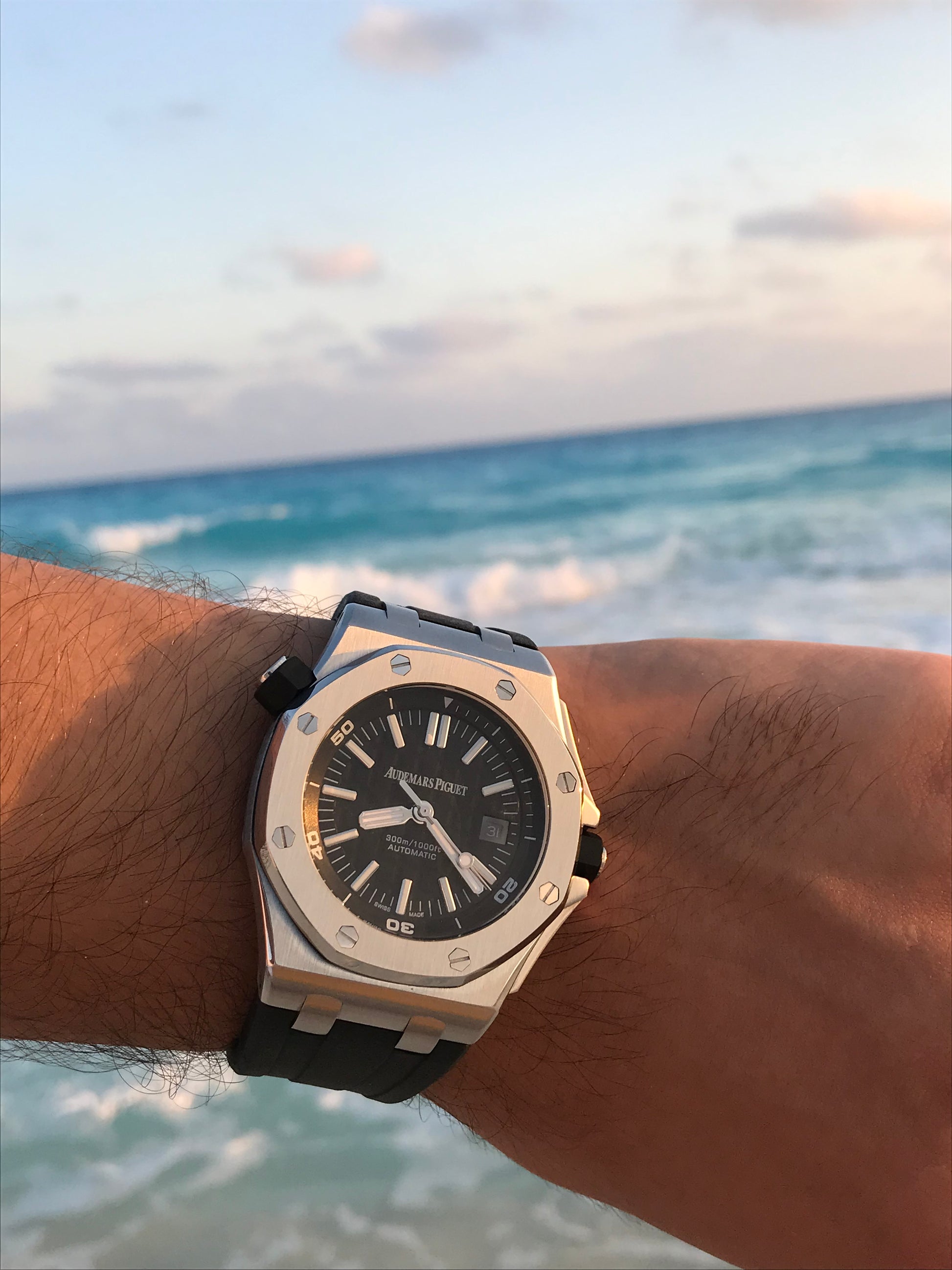 A close-up of a wrist wearing an Audemars Piguet Royal Oak watch with a black dial and black strap, set against a blurred beach and ocean background.
